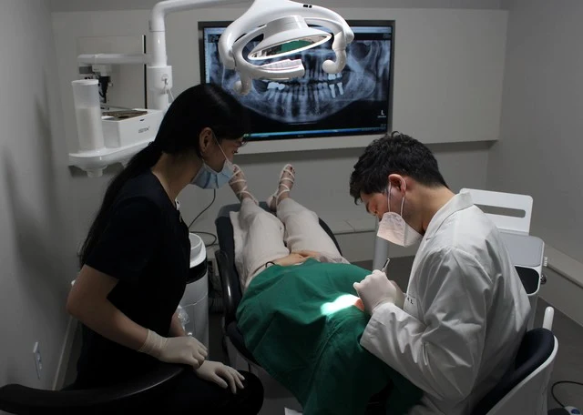 진료실에서 원장과 진료 보조 인력이 누운 환자의 임플란트 수술을 진행하는 모습. 뒤쪽 모니터에 파노라마 영상이 떠 있다.